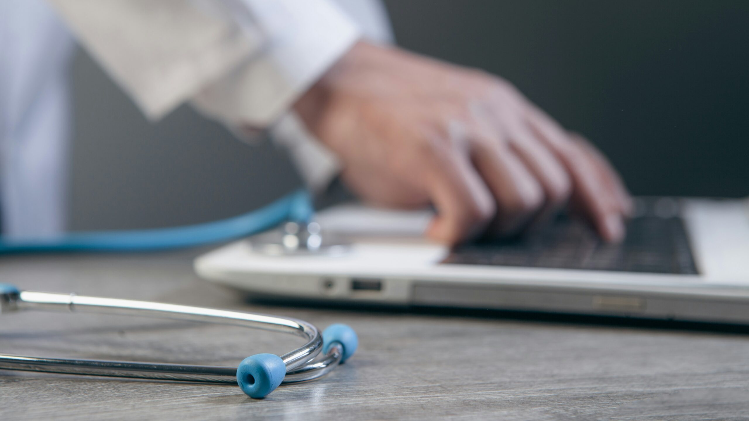 Doctor typing on a laptop with a stethoscope nearby.