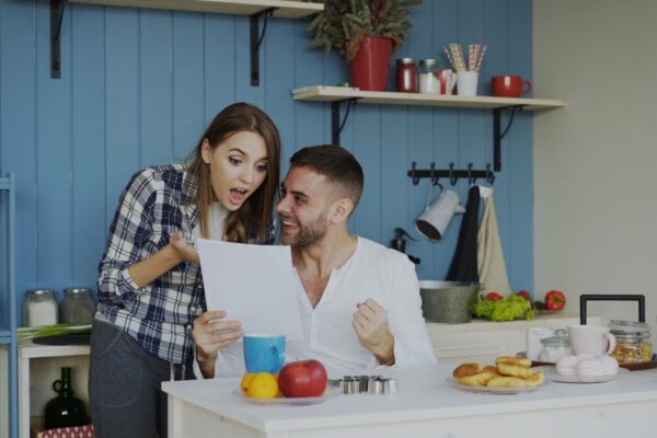 Attractive happy couple recieve good news unfolding letter in the kitchen while have breakfast