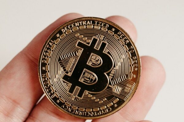 A close-up image of a Bitcoin coin held in a human hand against a white background.