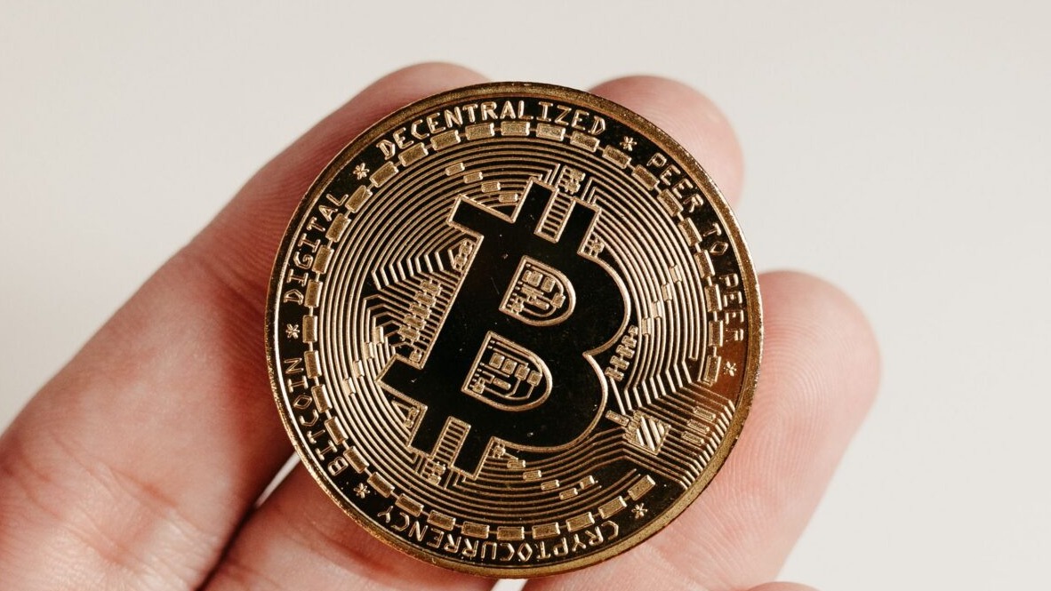 A close-up image of a Bitcoin coin held in a human hand against a white background.