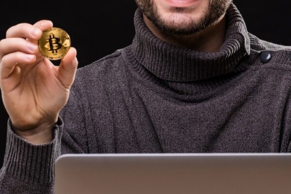 Portrait of a happy entrepreneur showing bitcoin while sitting at desk with laptop computer isolated over black