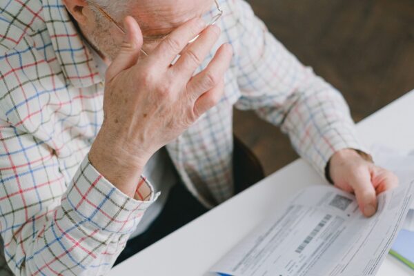Elderly man in patterned shirt reading and holding bills at a home table, appearing focused.