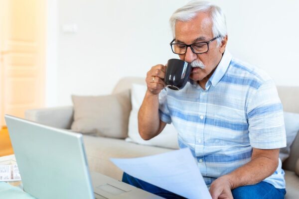 Focused greyhaired elderly man sit on couch reading bank notifications calculating domestic expenditures concentrated modern mature male consider financial paperwork pay bills on laptop online