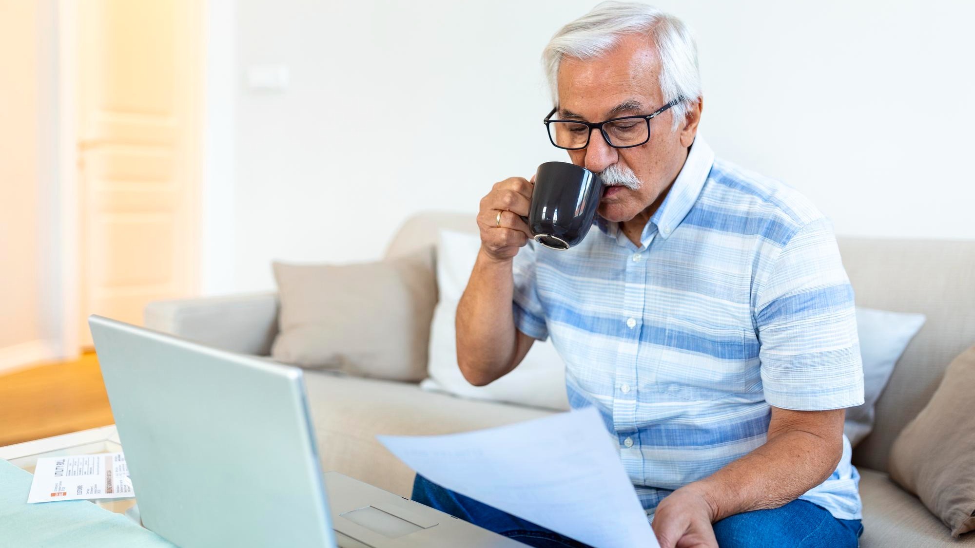 Focused greyhaired elderly man sit on couch reading bank notifications calculating domestic expenditures concentrated modern mature male consider financial paperwork pay bills on laptop online