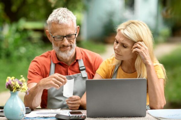 Stressed senior couple with laptop checking bills sitting at table in garden