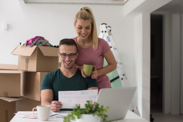 Young couple moving in a new home. Man and woman at the table using notebook laptop computer and plans with boxes around them