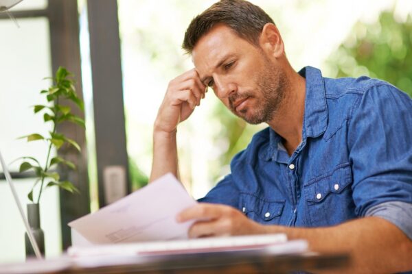 Stress documents and man in home for finance budget financial review and mortgage payment Thinking reading and frustrated person with paperwork for insurance investment and bills for debt