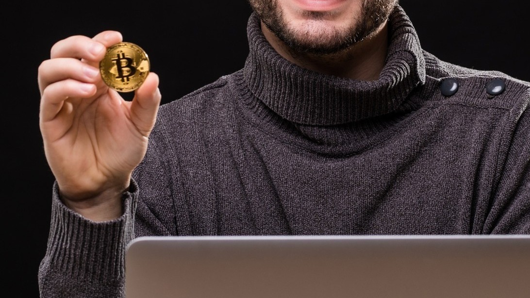 Portrait of a happy entrepreneur showing bitcoin while sitting at desk with laptop computer isolated over black