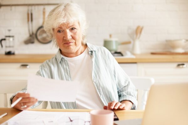 Elderly housewife with gray hair sitting in kitchen with open laptop and papers on table, having emotional frustrated facial expression, shocked with debt amount while paying domestic bills online