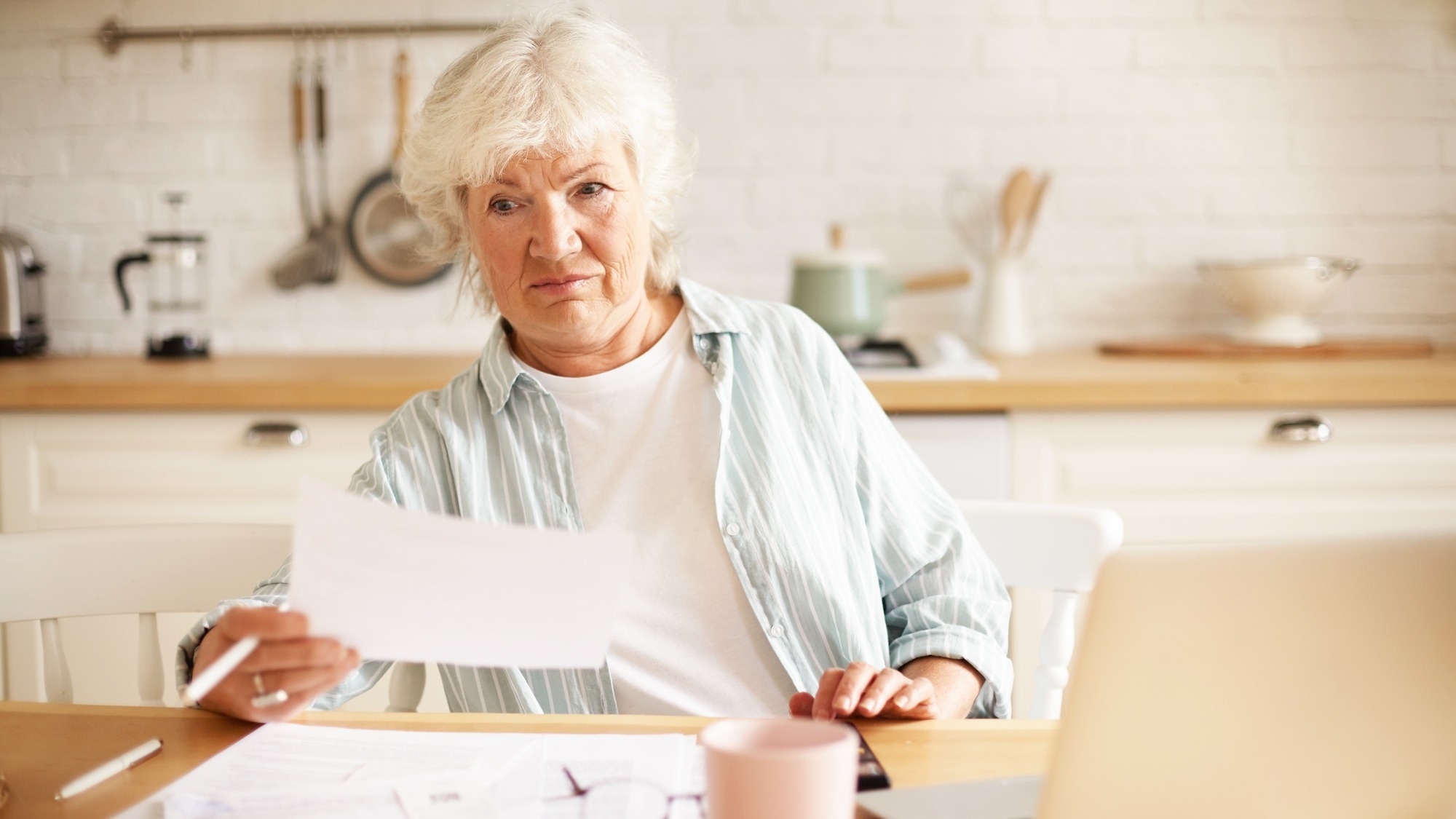 Elderly housewife with gray hair sitting in kitchen with open laptop and papers on table, having emotional frustrated facial expression, shocked with debt amount while paying domestic bills online