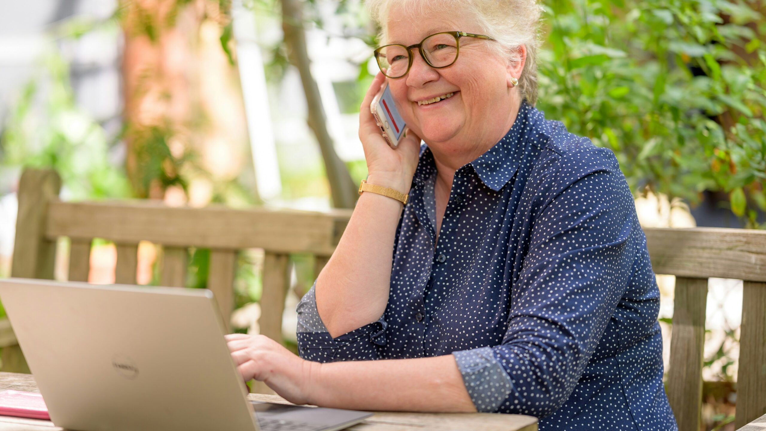 a woman on her phone while sitting at a table with a laptop