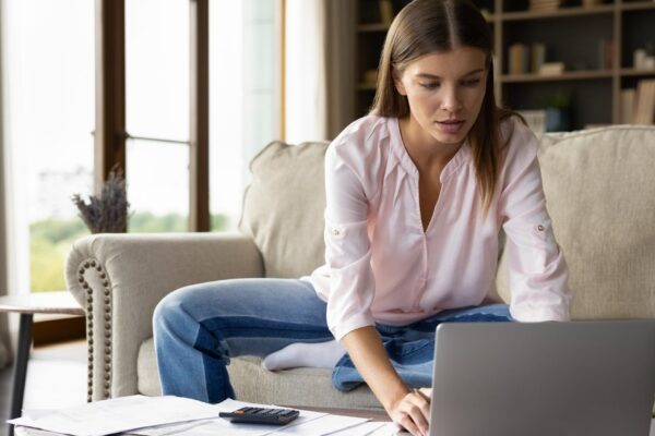 Focused young homeowner girl using payment banking financial online application on laptop computer checking domestic paper documents paying bills calculating taxes insurance mortgage fees