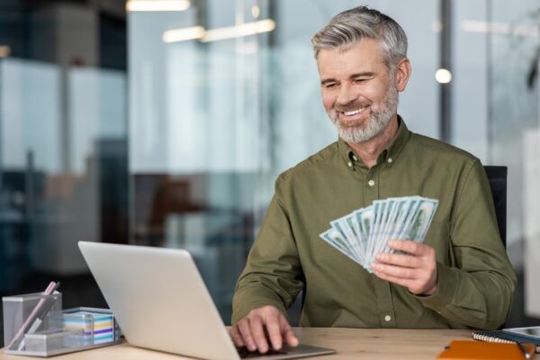 Mature businessman smiling holding a fan of dollar bills while using his laptop at an office desk
