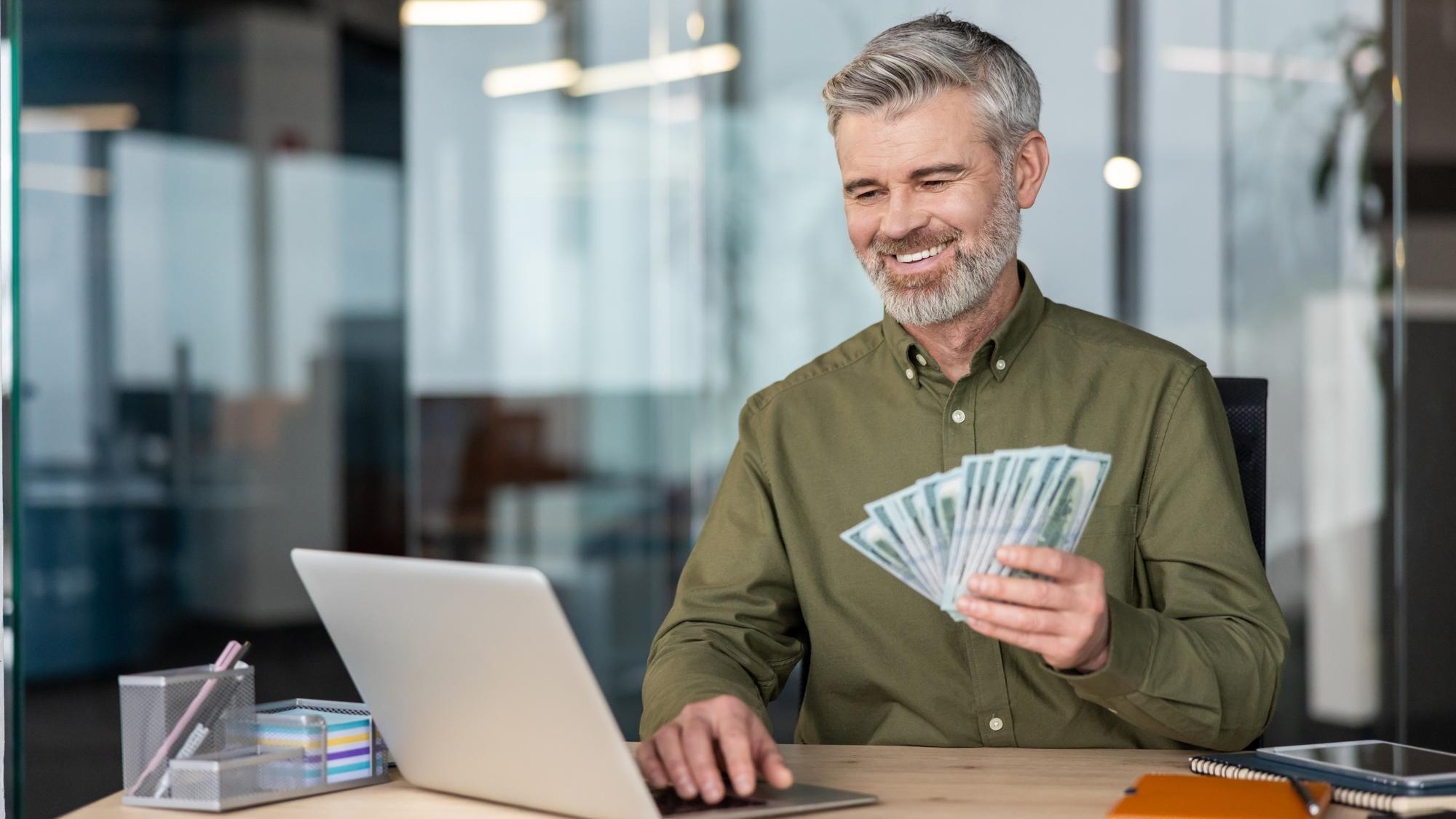 Mature businessman smiling holding a fan of dollar bills while using his laptop at an office desk