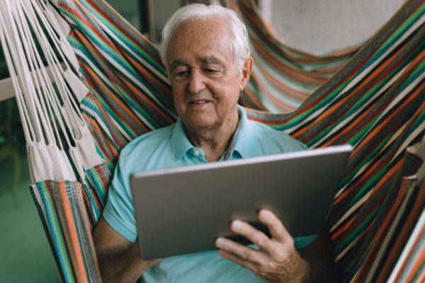 Elderly man enjoying leisure time in a hammock while using a tablet indoors.