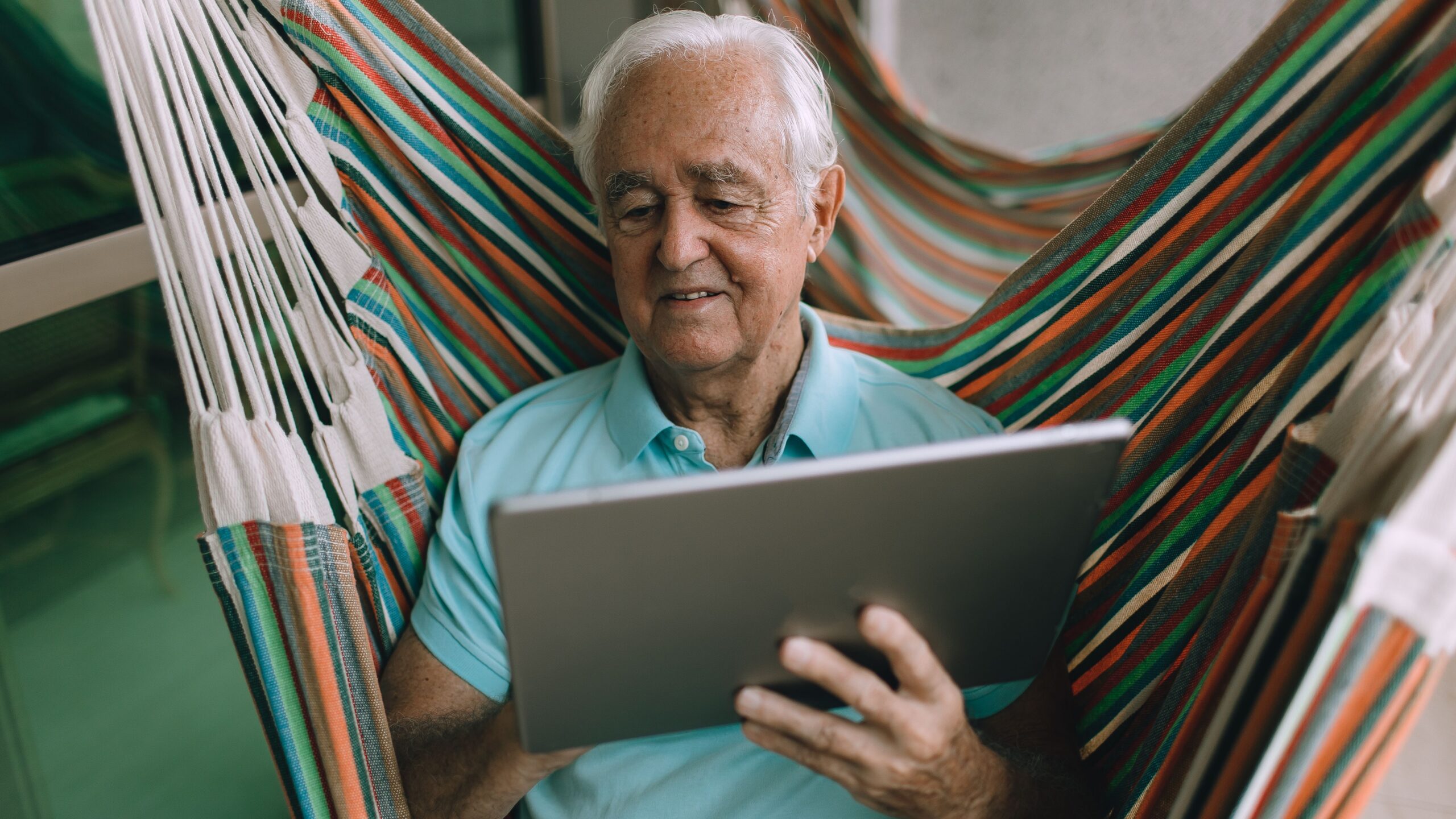 Elderly man enjoying leisure time in a hammock while using a tablet indoors.