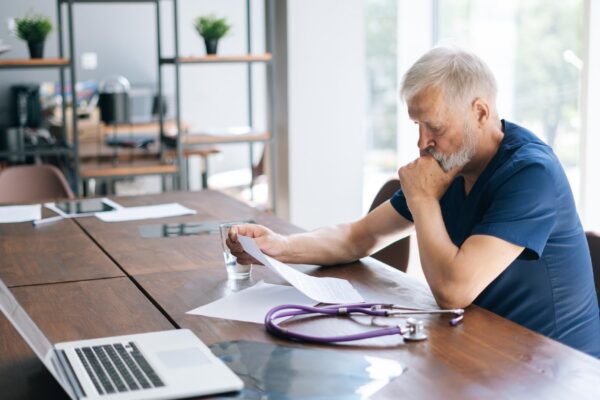 Focused thoughtful gray-haired mature man doctor studying medical report of patient, working with paper documents sitting at desk in office room in medical clinic.