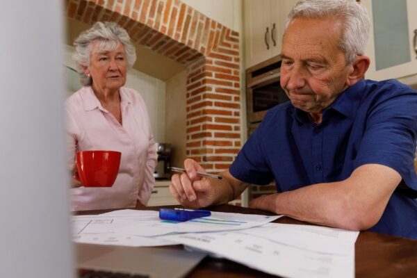 Senior caucasian couple spending time at home together, sitting in kitchen, the man looking at paperwork and the woman holding a cup. isolating during coronavirus covid 19 quarantine lockdown.