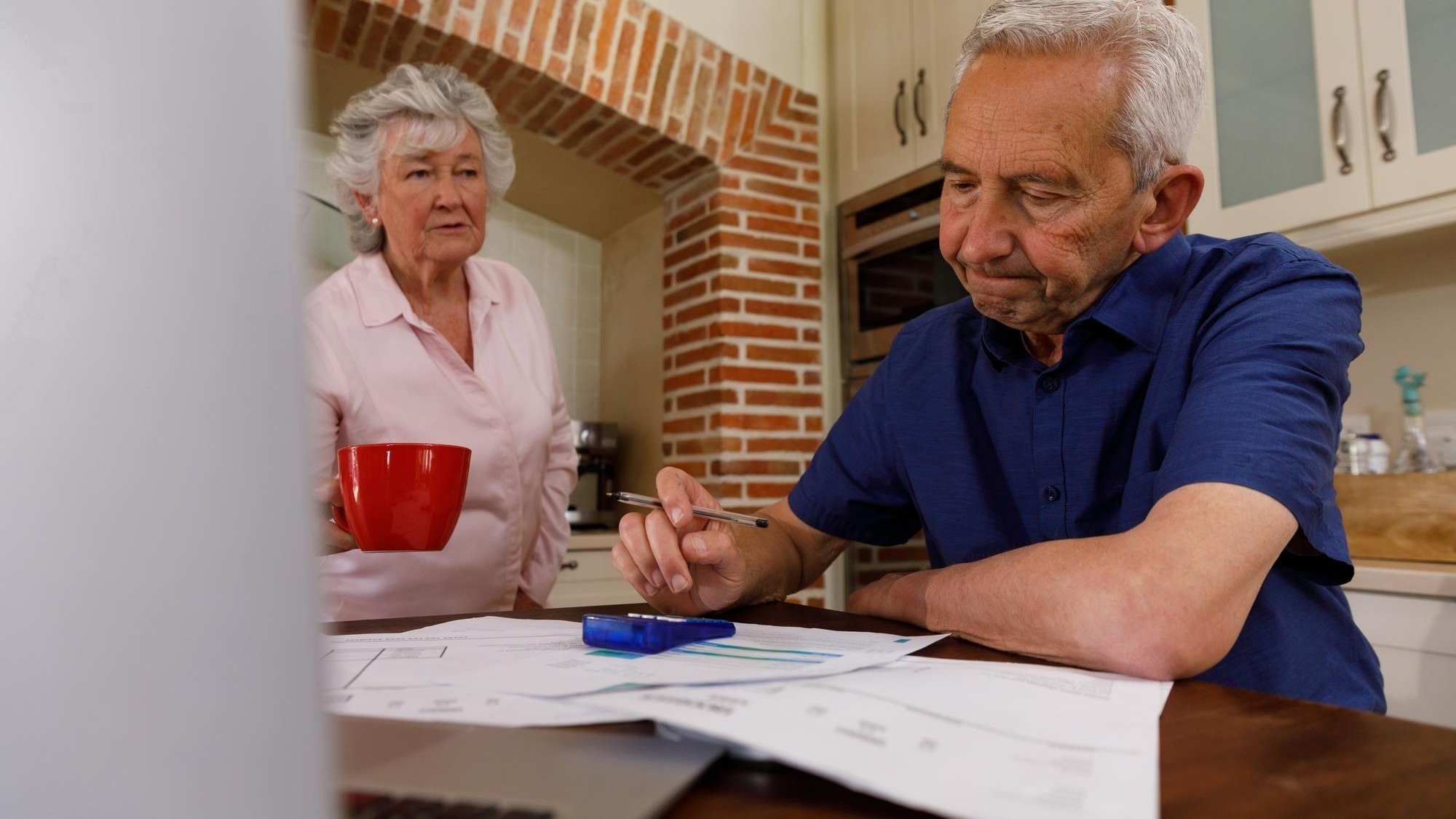 Senior caucasian couple spending time at home together, sitting in kitchen, the man looking at paperwork and the woman holding a cup. isolating during coronavirus covid 19 quarantine lockdown.
