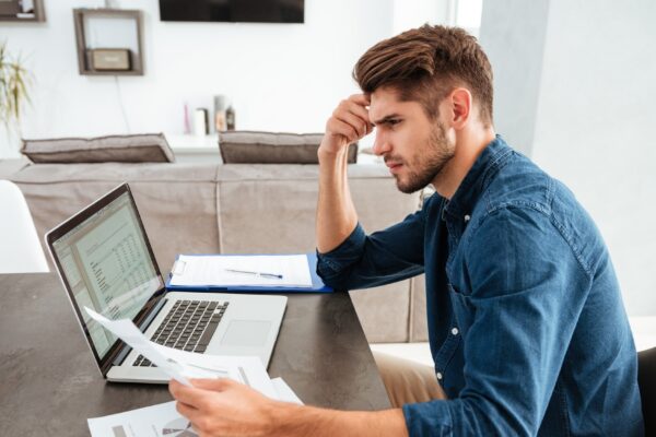 Serious concentrated man working at laptop and sitting at the table while looking at the papers and holding his head with hand