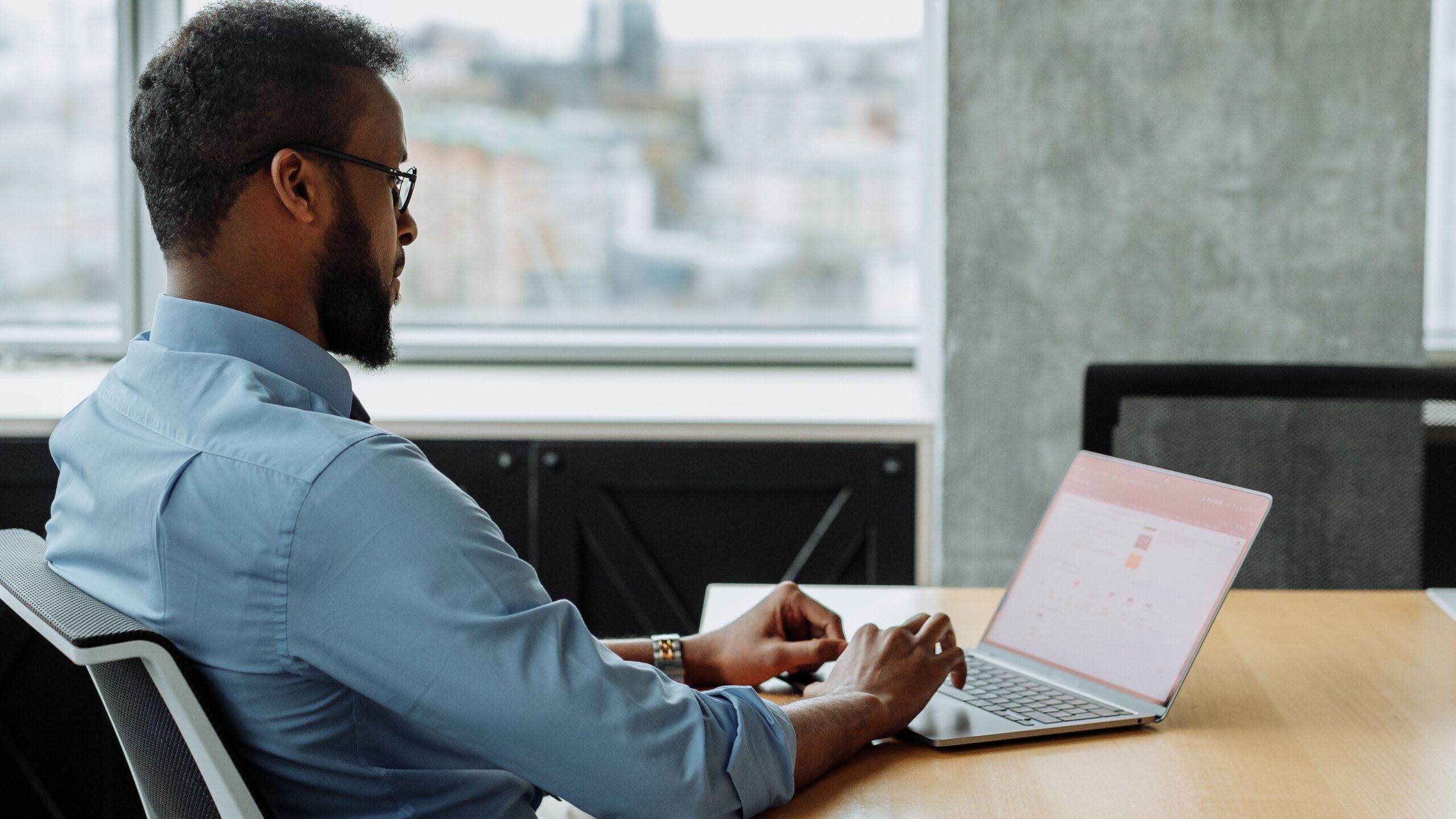 Black man working on laptop in modern office with city view, focused on technology and productivity.