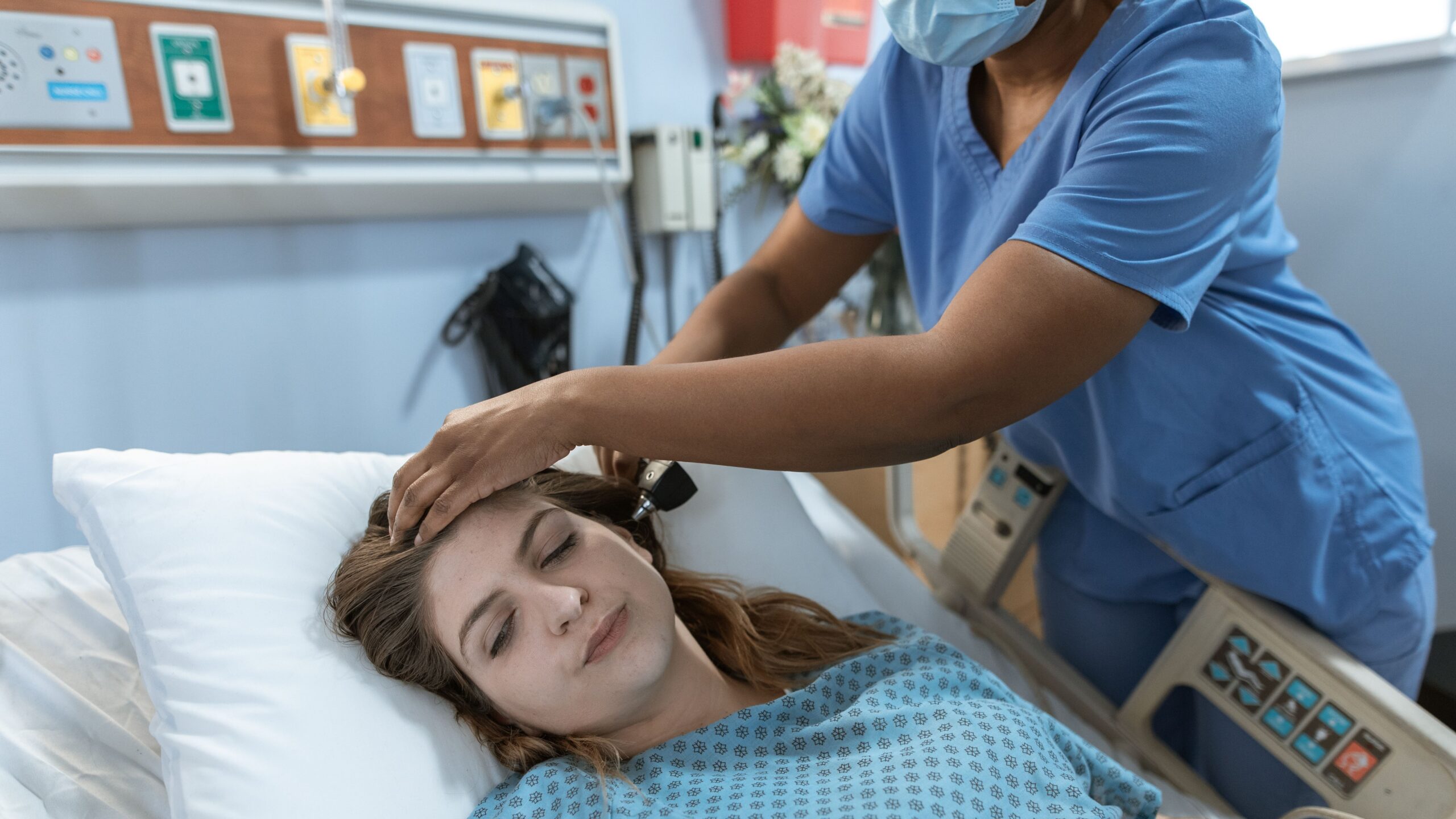Crop African American female doctor with professional equipment doing examination of ear of woman lying on bed in hospital ward