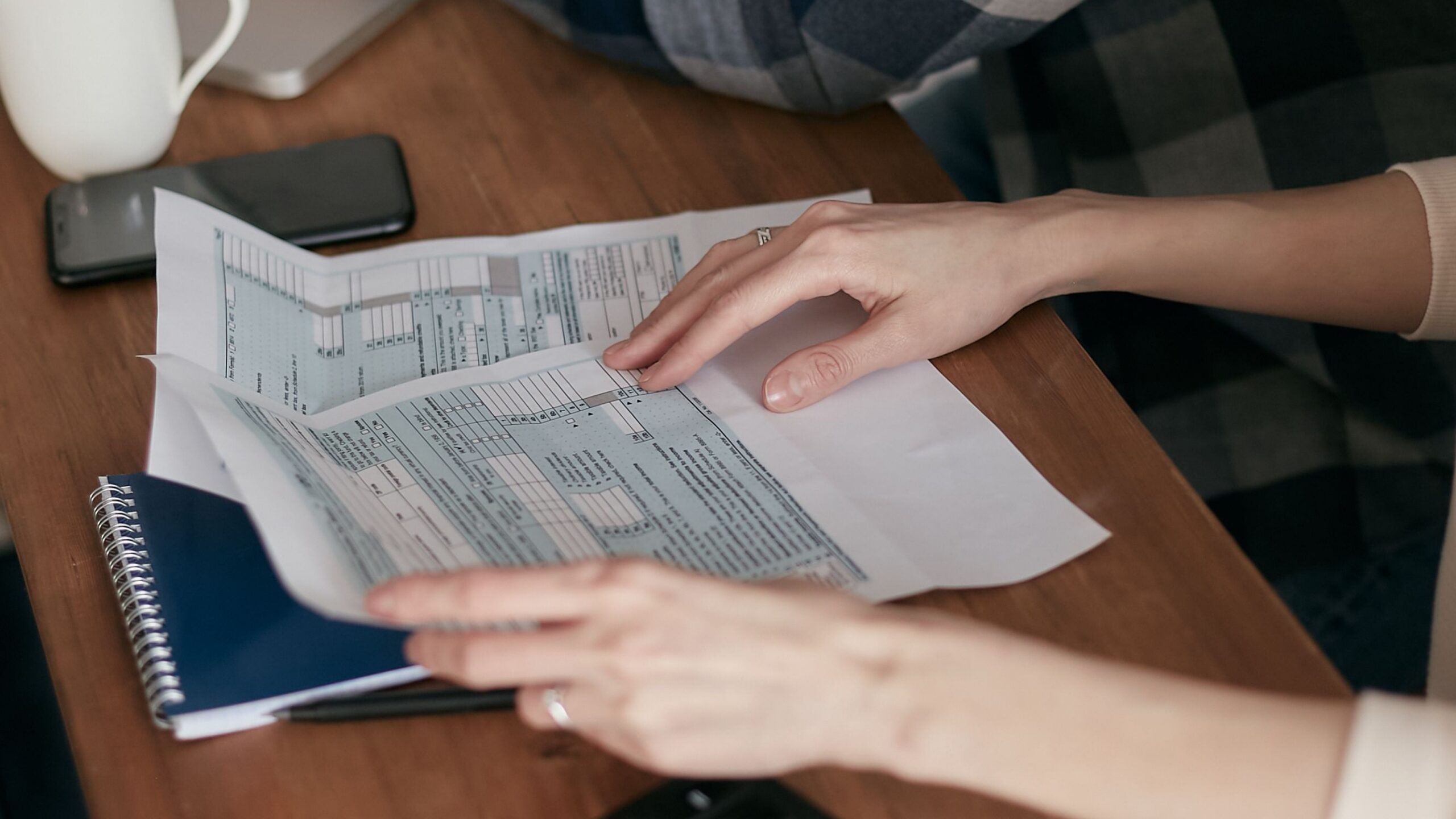 Close-up of hands organizing tax forms on a desk with a calculator, laptop, and notebook.