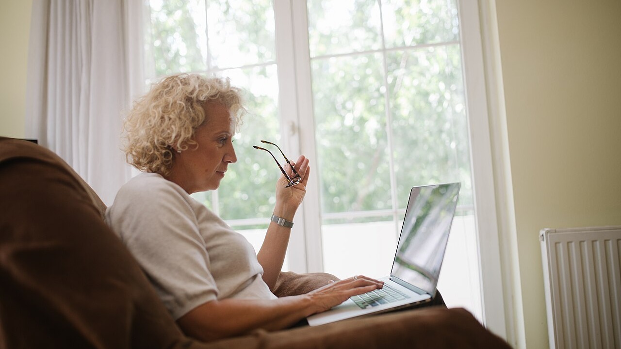 Elderly woman sitting in an armchair and holding her glasses while typing on a laptop.