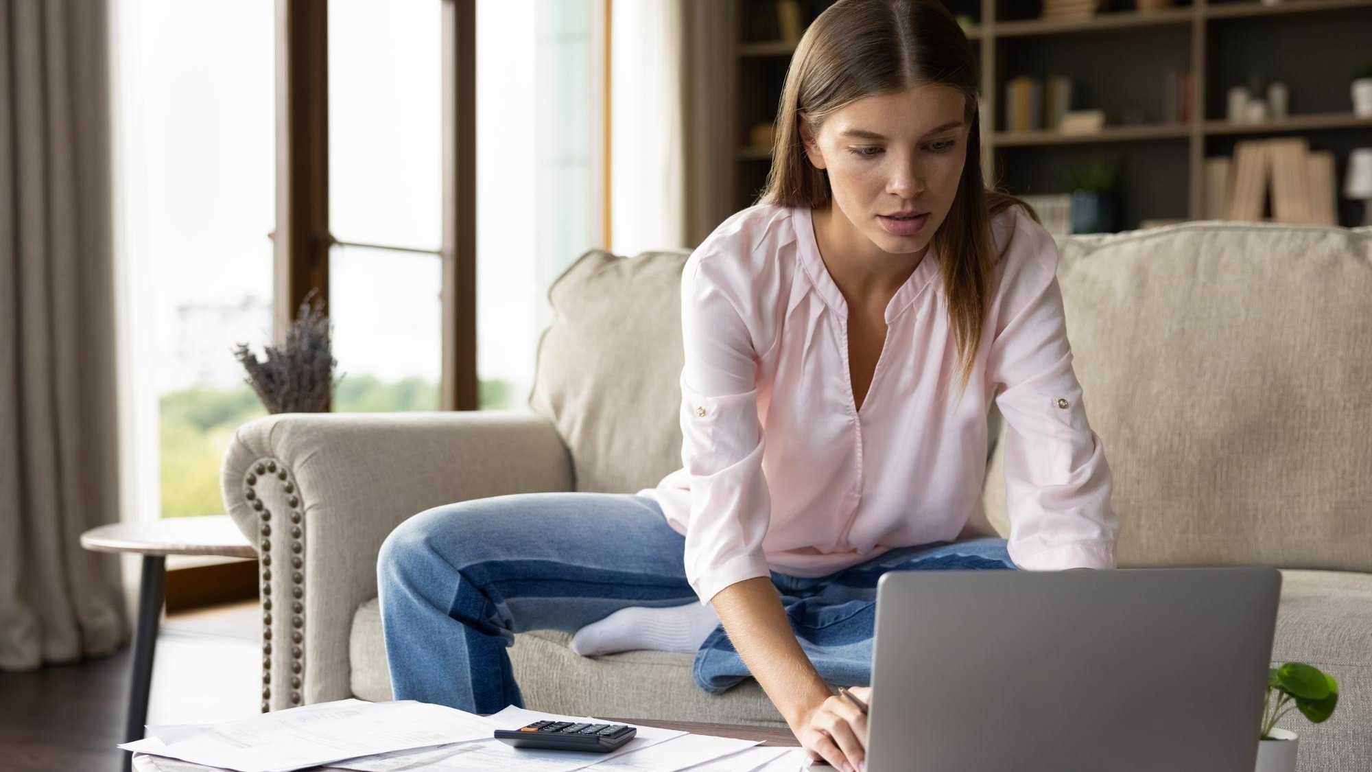Focused young homeowner girl using payment banking financial online application on laptop computer checking domestic paper documents paying bills calculating taxes insurance mortgage fees