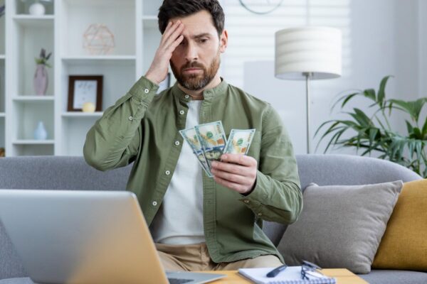 Worried young man sitting at home on the sofa in front of the table with a laptop holding his head with his hand and looking sadly at the cash money in his hand