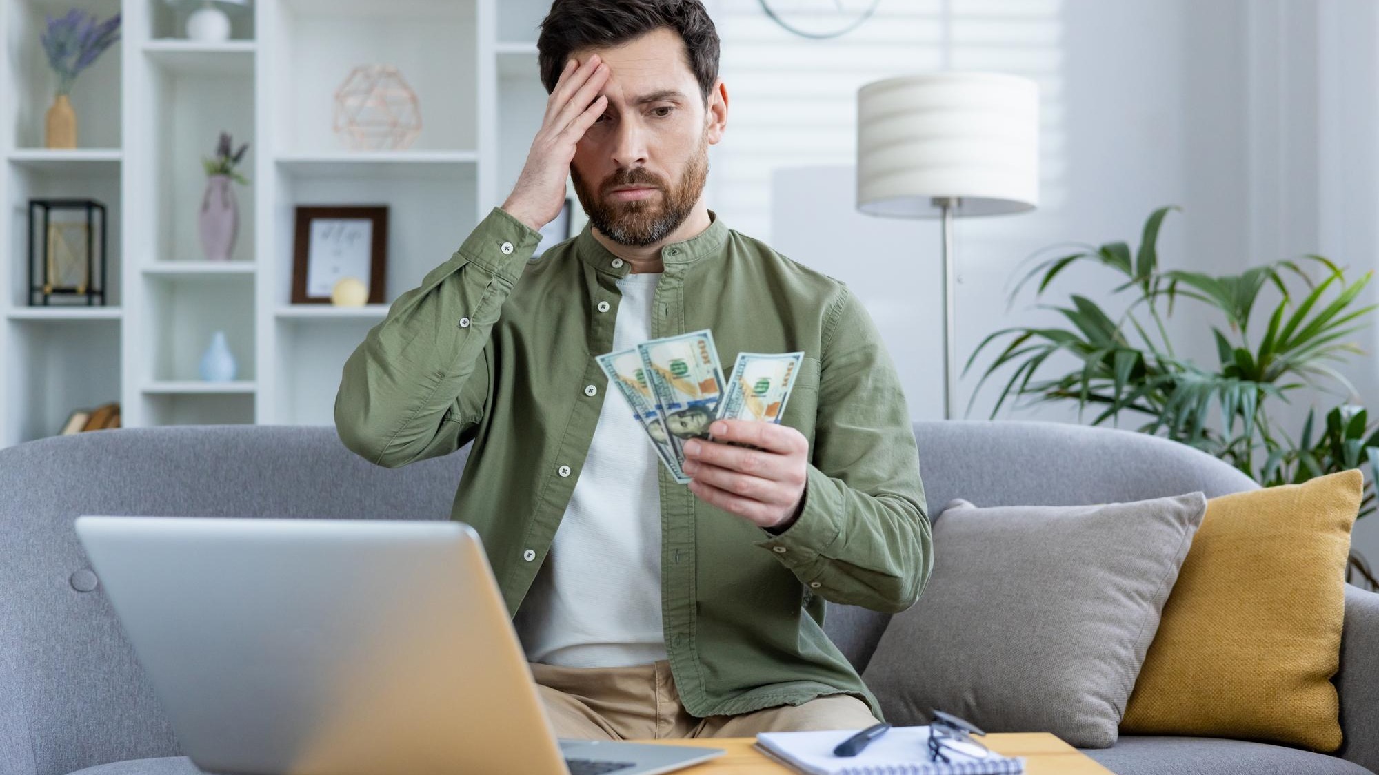 Worried young man sitting at home on the sofa in front of the table with a laptop holding his head with his hand and looking sadly at the cash money in his hand