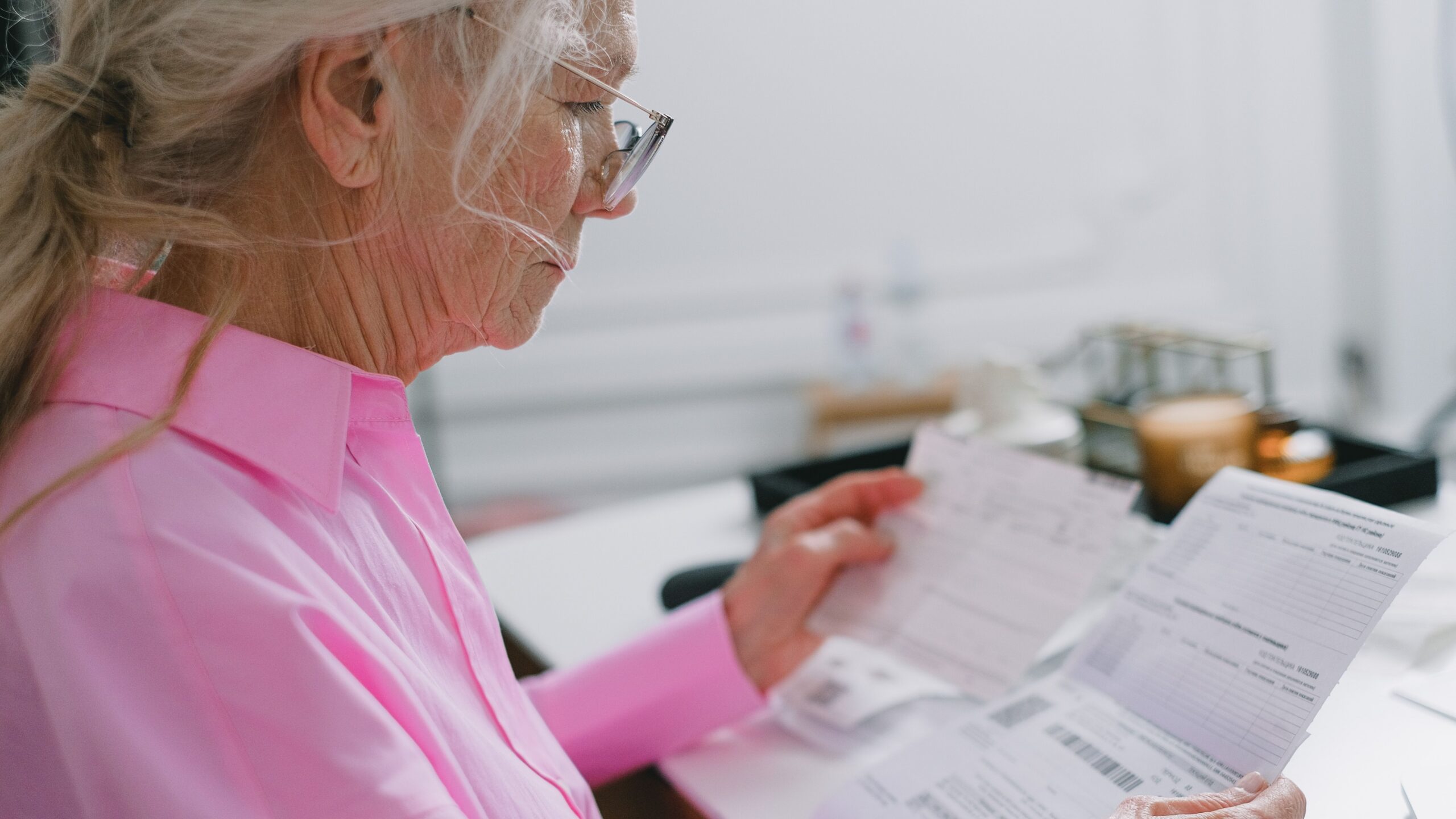 Elderly woman in a pink shirt reviewing paperwork at home.