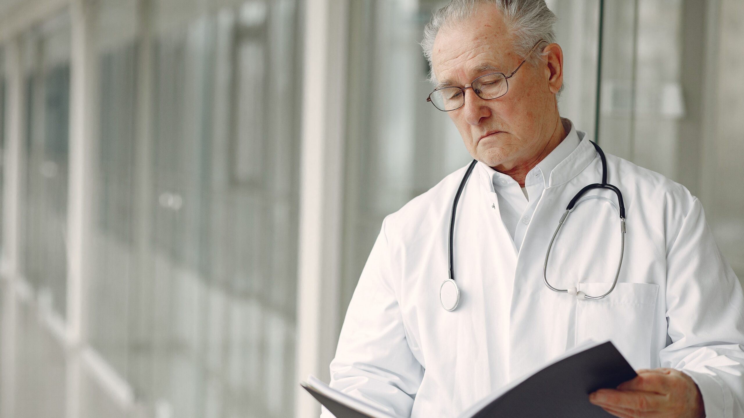 Crop concentrated professional senior male physician in white robe with stethoscope standing in modern hospital hallway and reading medical case records in daylight