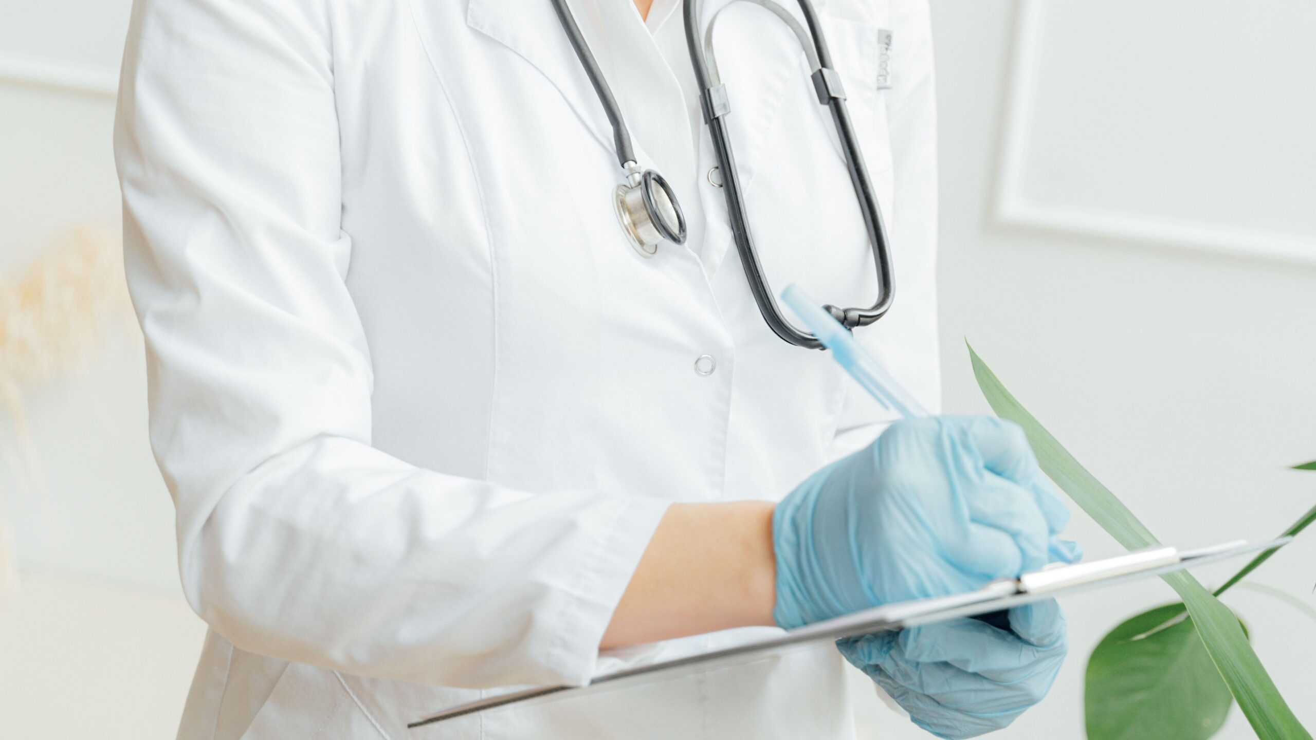 Female doctor with eyeglasses taking notes in a hospital, wearing a stethoscope and gloves.