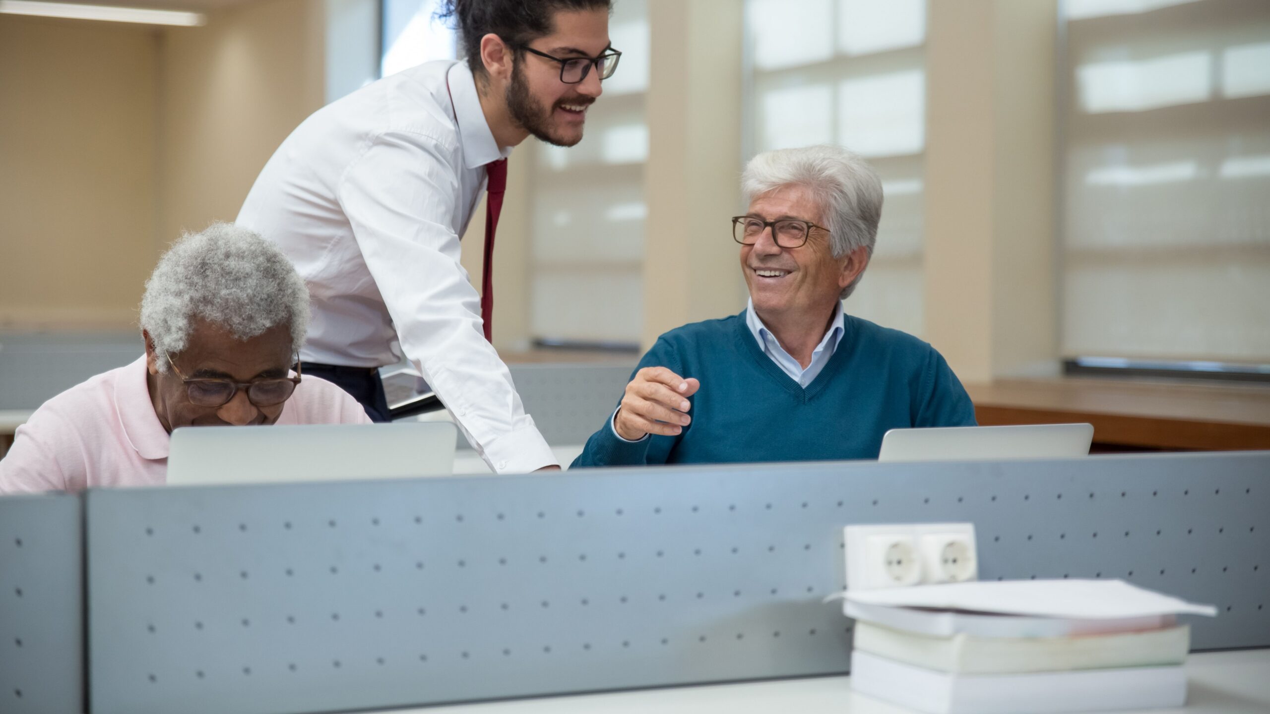 A classroom scene in Portugal where seniors learn computer skills from a young instructor. Engaged and smiling.