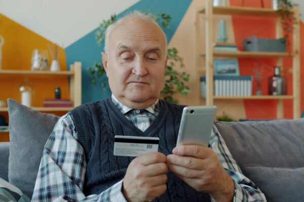 Elderly man holding credit card and smartphone for online shopping.