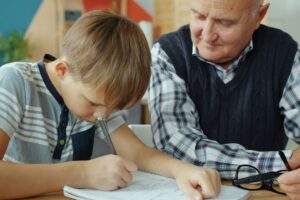 Grandfather helps grandson with homework at table.