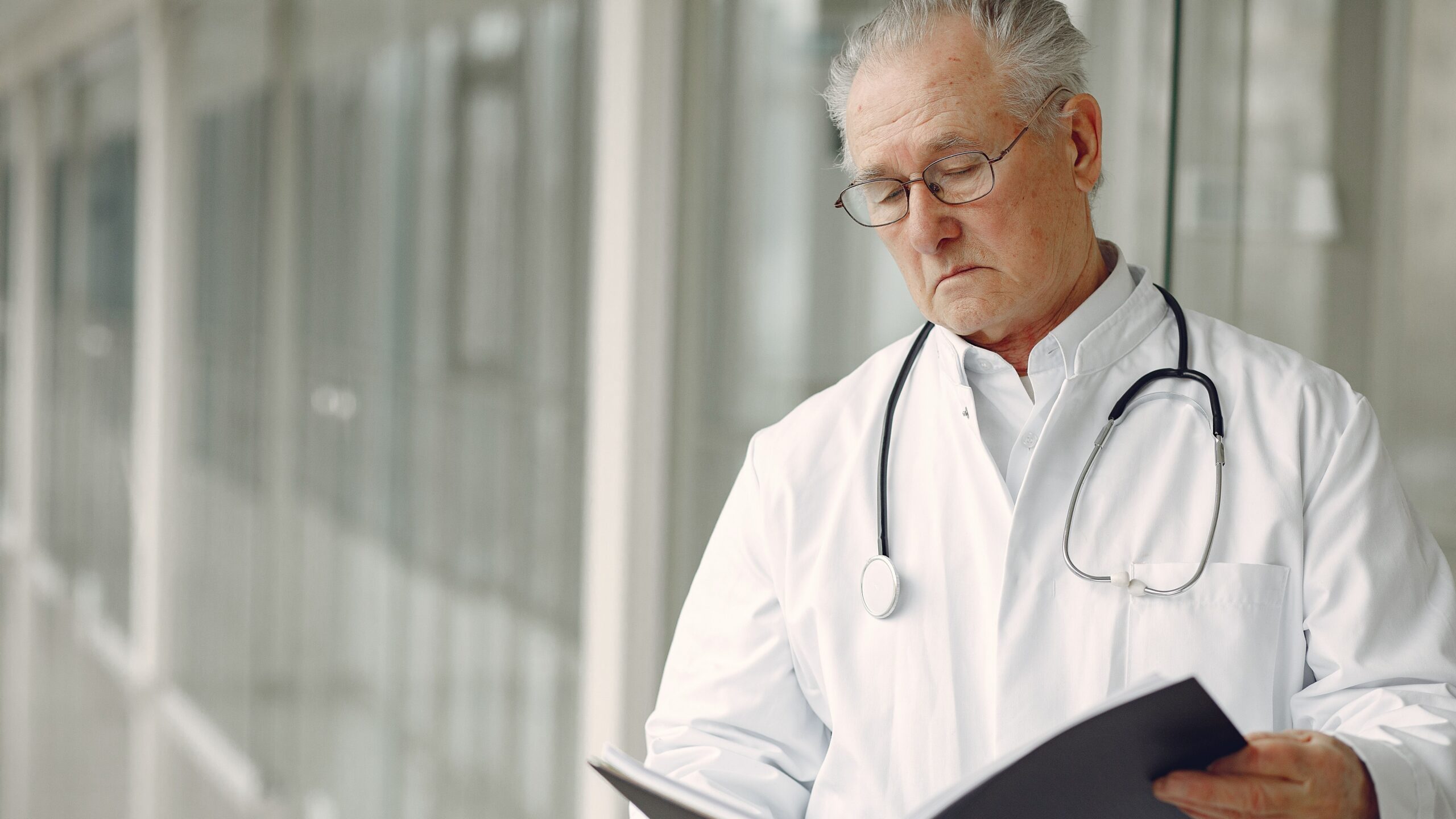 Crop concentrated professional senior male physician in white robe with stethoscope standing in modern hospital hallway and reading medical case records in daylight