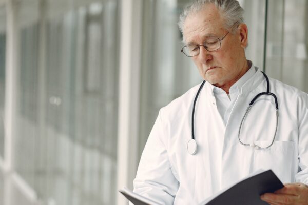 Crop concentrated professional senior male physician in white robe with stethoscope standing in modern hospital hallway and reading medical case records in daylight