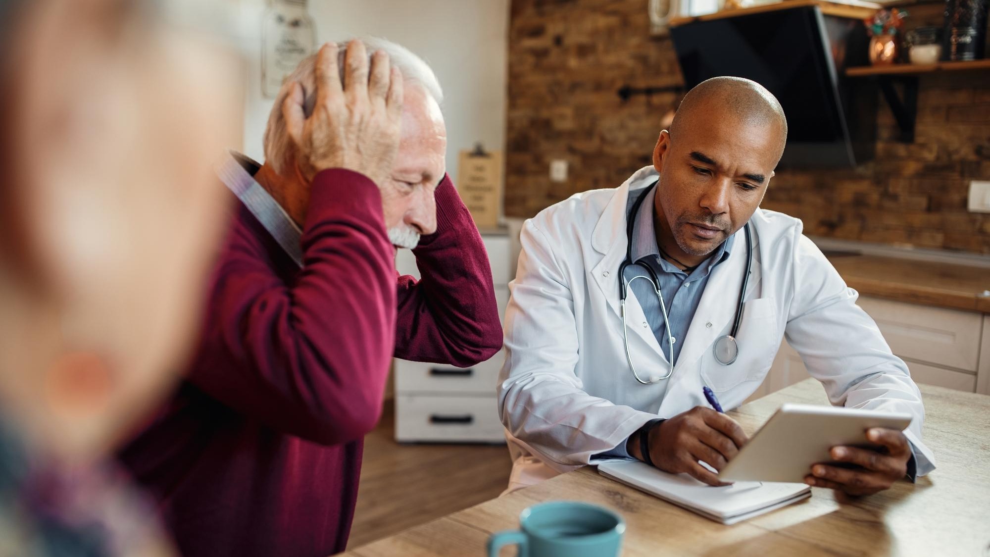 Black doctor using touchpad and taking notes during a visit at senior man's home