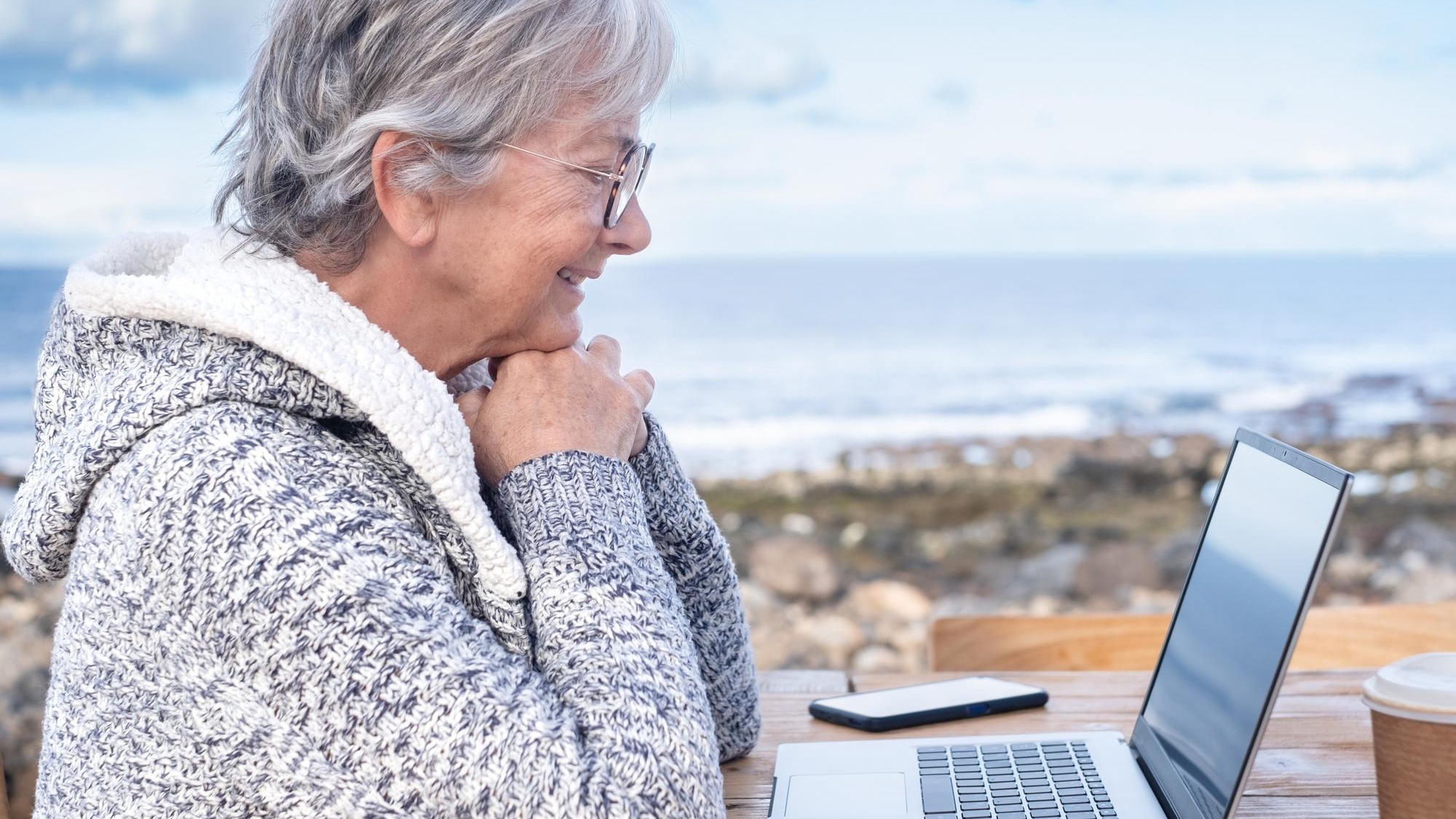 Smiling attractive mature senior adult professional business woman in remote working on laptop computer by the beach wearing eyeglasses sitting at the wooden table looking at computer screen