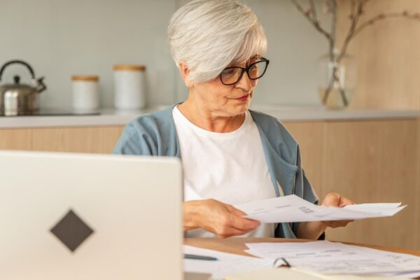 Middle aged senior woman sit with laptop and paper document pensive older mature lady reading paper