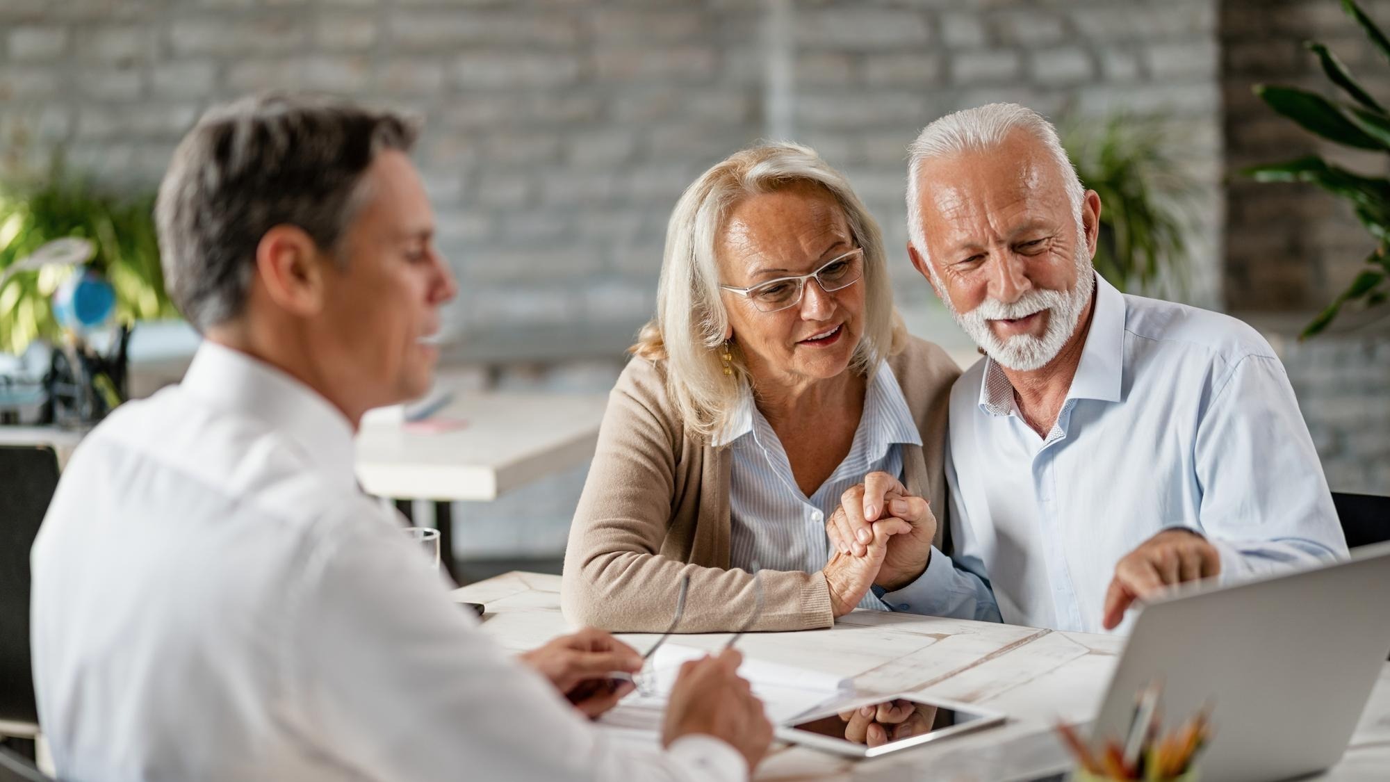 Happy senior couple holding hands and using laptop while having a meeting with financial advisor in the office Senior man is pointing at something on laptop