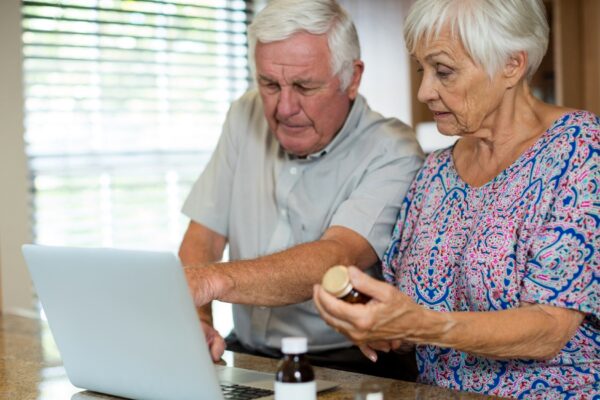 Senior couple using laptop and holding pill bottle in kitchen at home