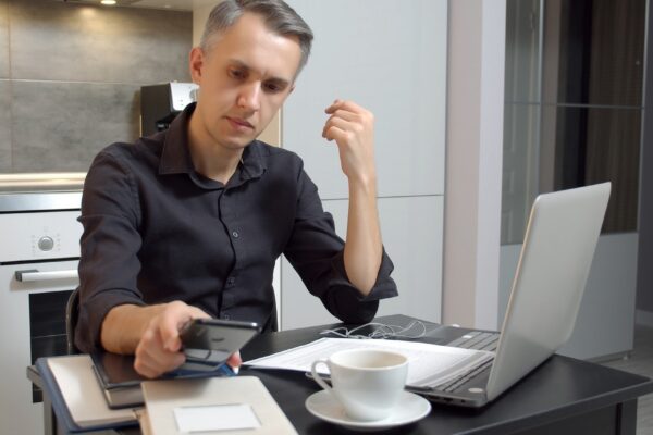 Focused young man working on laptop and using smartphone while sitting in kitchen at home