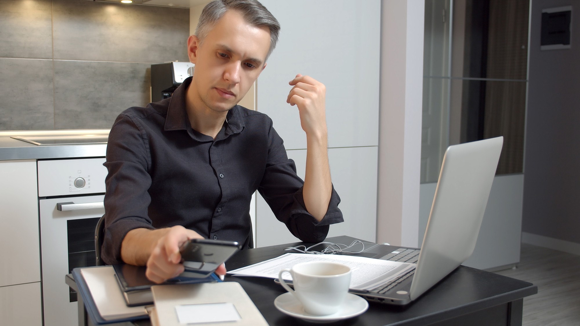 Focused young man working on laptop and using smartphone while sitting in kitchen at home