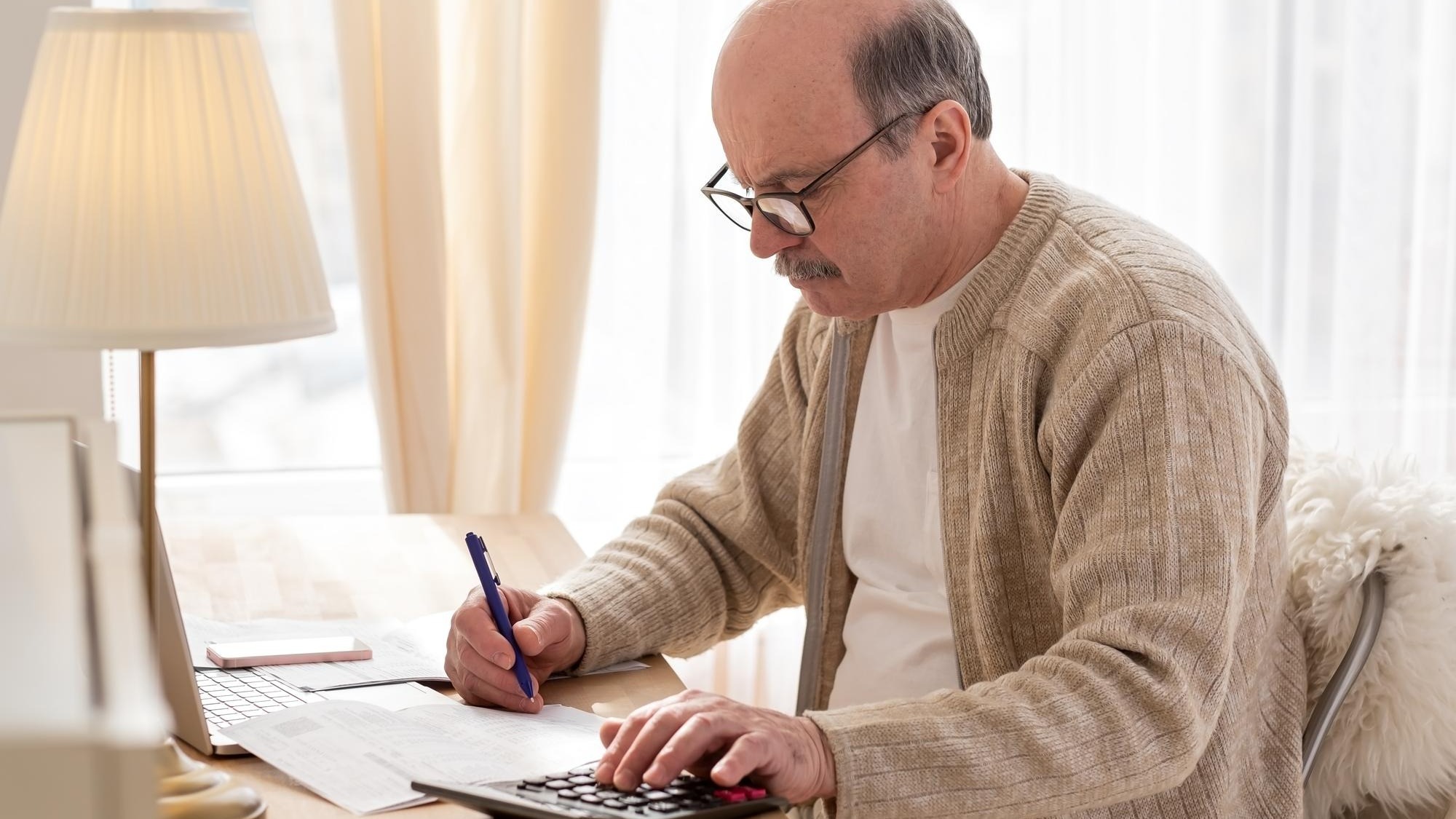 Senior man sitting with paperwork and using calculator while counting money