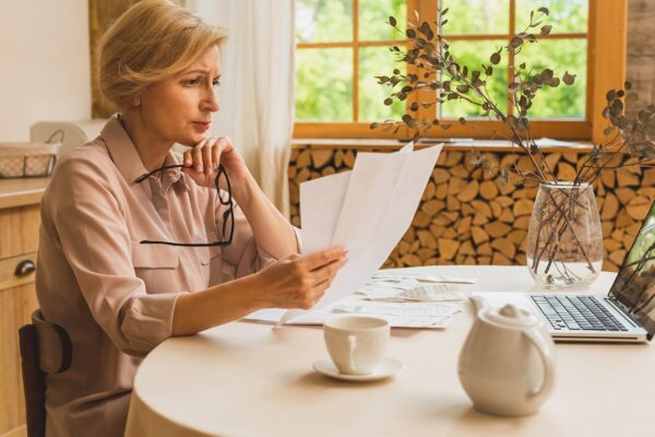 Middle aged mature senior woman holding paper bill or letter using laptop computer at home for making online payments on website, calculating financial taxes fee cost, reviewing bank account.