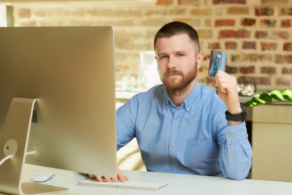 A man with a beard in a blue shirt posing with his credit card in front of the computer at home