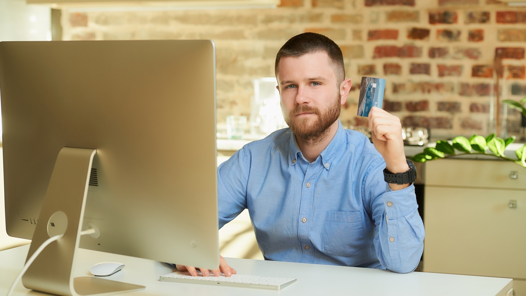 A man with a beard in a blue shirt posing with his credit card in front of the computer at home